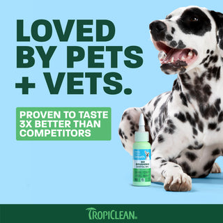 White and black spotted dog next to TropiClean Fresh Breath pet toothpaste bottle with green label on blue background.